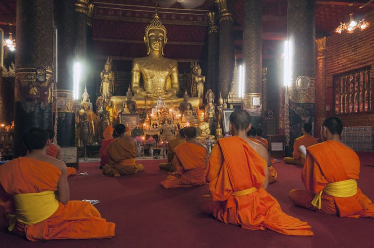 Monks dressed in saffron robes seated in rows before a large golden Buddha icon.